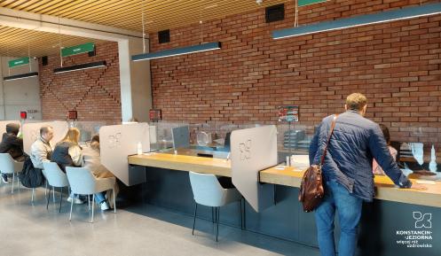 Wnętrze urzędu z kilkoma stanowiskami obsługi, przy których siedzą lub stoją petenci, a nad nimi wiszą zielone tablice informacyjne w języku polskim.