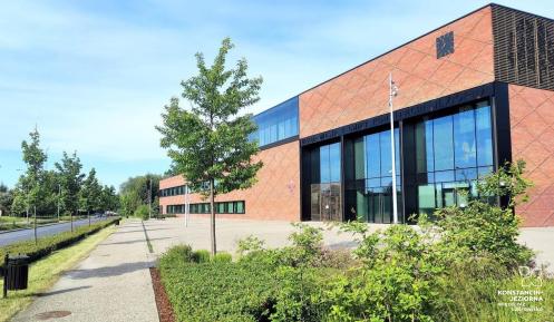 Nowoczesny budynek Urzędu Miasta i Gminy Konstancin-Jeziorna z czerwonej cegły, otoczony zielenią i ścieżką pieszą w słoneczny dzień.