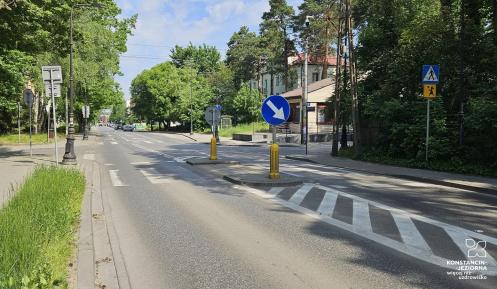 Ulica z wysepką rozdzielającą pasy ruchu, znakami drogowymi oraz przejściem dla pieszych, otoczona drzewami i budynkami.
