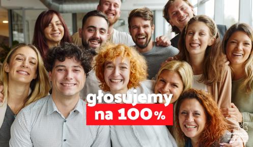 Grupa uśmiechniętych ludzi pozuje razem do zdjęcia, a na pierwszym planie widnieje hasło „głosujemy na 100%” na czerwono-białym tle.
