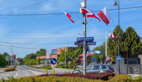 Rondo ozdobione polskimi flagami, otoczone znakami drogowymi i zielenią, znajduje się przy ulicy Warszawskiej w słoneczny dzień.