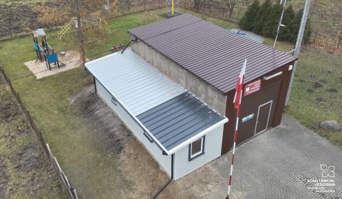 Widok z lotu ptaka na budynek Ochotniczej Straży Pożarnej w Słomczynie z placem zabaw w tle i maszt z polską flagą przed wejściem.