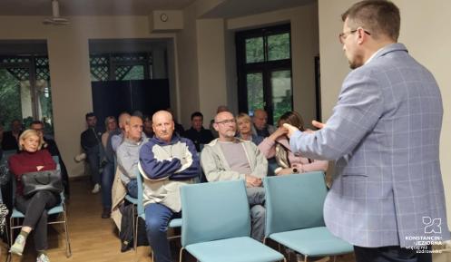 Mężczyzna w marynarce przemawia do zgromadzonych mieszkańców siedzących przy stołach, podczas spotkania w świetlicy wiejskiej.