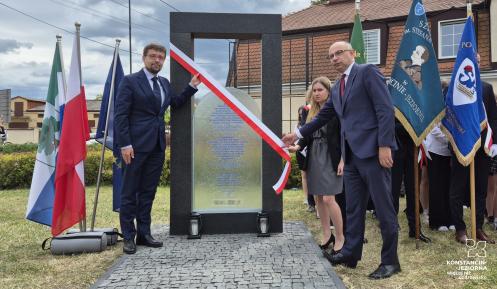 Uroczyste odsłonięcie pomnika upamiętniającego, z udziałem oficjeli i pocztów sztandarowych, odbywa się przy asyście flag narodowych i lokalnych.