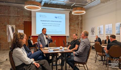 grupa ludzi siedzi przy stole, za nimi widać planszę z tytułem warsztaty strategiczne diagnoza otoczenia zewnętrznego