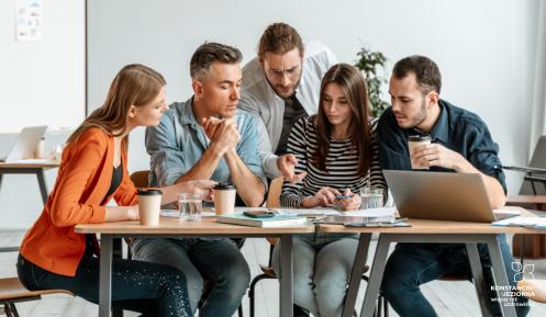 Grupa ludzi siedzi przy stole - rozmawiają o projekcie. Na stole laptop, kubki z kawą, dokumenty. W tle tablica do pisania.
