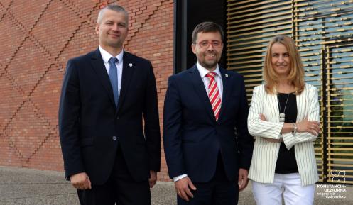 Na zdjęciu trzech uśmiechniętych przedstawicieli samorządu gminy Konstancin-Jeziorna pozuje przed nowoczesnym budynkiem z ceglaną i przeszkloną elewacją.