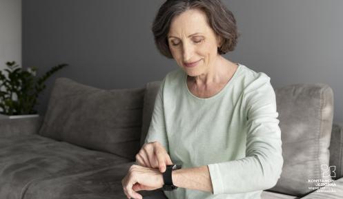 Starsza kobieta siedzi na sofie i uśmiechając się, obsługuje opaskę sportową lub smartwatch na swoim nadgarstku.