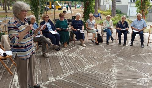 Grupa osób siedzi w altanie, jedna osoba czyta książkę.