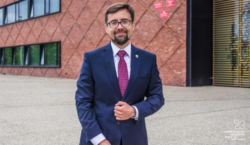 uśmiechnięty burmistrz w granatowym garniturze stoi na tle urzędu miasta i gminy Konstancin-Jeziorna