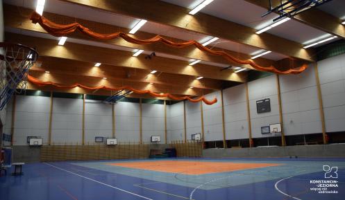 Wnętrze dużej, nowoczesnej hali sportowej z wieloma koszami do koszykówki, niebieską podłogą i pomarańczowymi siatkami pod sufitem.