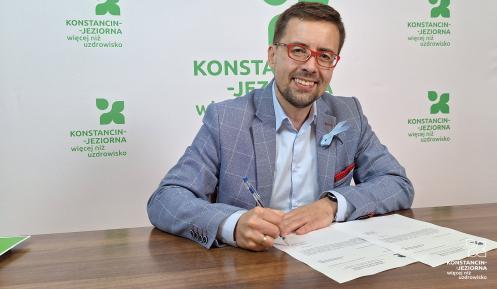 Uśmiechnięty mężczyzna w okularach i eleganckiej marynarce siedzi przy biurku i podpisuje dokumenty na tle banneru promocyjnego miasta.