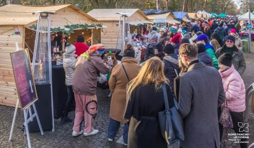 Ludzie spacerują między drewnianymi stoiskami z ozdobami i świątecznymi produktami podczas jarmarku w parku otoczonym drzewami.