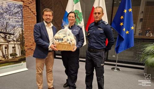 trzy osoby stoją na tle flag trzymając kosz z białymi maskotkami