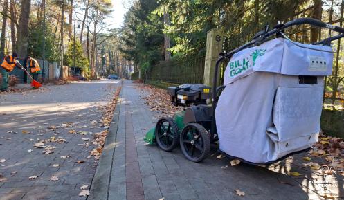 Odkurzacz do liści na ulicy zasypanej liściami, w tle dwoje pracowników z grabiami porządkuje liście.