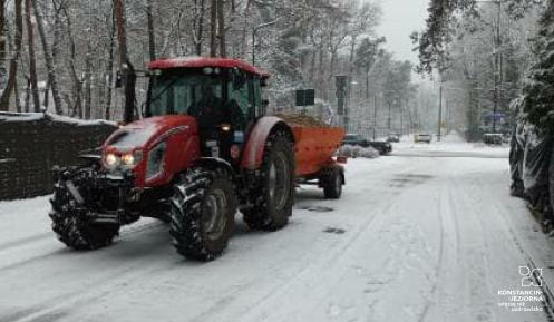 Na ośnieżonej ulicy jedzie czerwony traktor z posypywarką, wykonując zimowe prace utrzymaniowe w otoczeniu drzew i zaparkowanych samochodów.