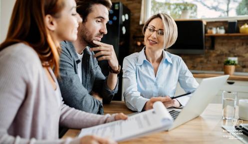 Trzy osoby siedzą przed komputerem, na stole leżą dokumenty, sprawdzają coś