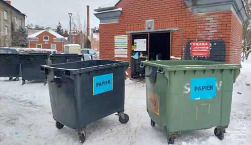 Altana śmietnikowa na osiedlu zimą, przed którą stoją przepełnione pojemniki na papier, ustawione na zaśnieżonym placu.
