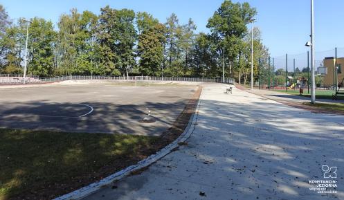 Szkolne boisko z asfaltową nawierzchnią i bramkami, położone obok budynku szkoły, otoczone zielenią i oświetleniem.