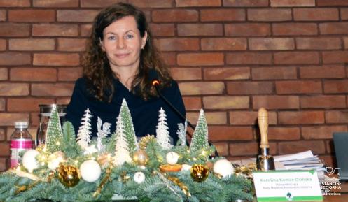 Uśmiechnięta kobieta za stołem prezydialnym, na tle ściany z czerwonej cegły.