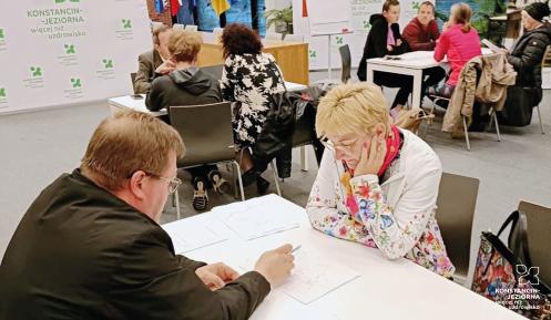 Mieszkańcy uczestniczą w warsztatach konsultacyjnych w sali urzędu, którzy przy stolikach omawiają mapy i dokumenty z przedstawicielami gminy.