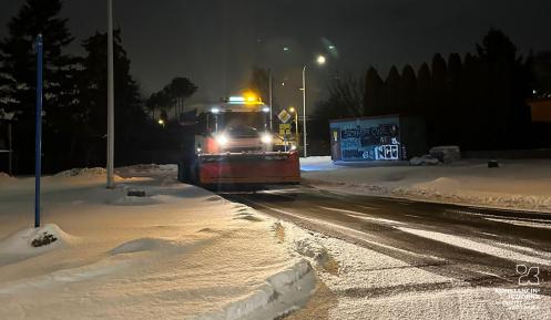Pług śnieżny odśnieżający zaśnieżoną jezdnię, oświetloną latarniami ulicznymi, z widocznymi śladami przejazdu na mokrym asfalcie.