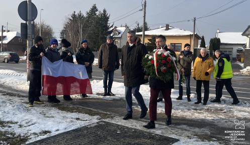 Grupa osób podczas zimowej uroczystości składa kwiaty pod pomnikiem, jedna z nich niesie wieniec, a w tle widoczna jest flaga Polski oraz zabudowa jednorodzinna przy drodze.