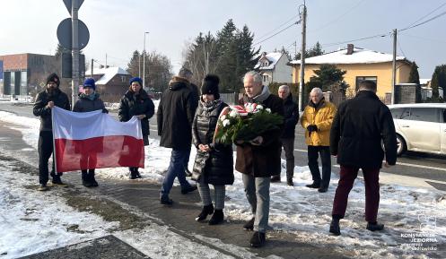 Grupa osób podczas zimowej uroczystości składa kwiaty pod pomnikiem, jedna z nich niesie wieniec, a w tle widoczna jest flaga Polski oraz zabudowa jednorodzinna przy drodze.
