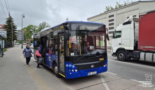Niebieski autobus komunikacji miejskiej linii L55 stoi na przystanku, do którego wsiadają pasażerowie, a obok na jezdni przejeżdża samochód ciężarowy.
