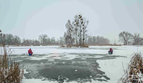 Dwóch wędkarzy siedzi na zamarzniętym stawie i łowi ryby w przeręblach, mimo cienkiej i nierównej tafli lodu pokrytej śniegiem.