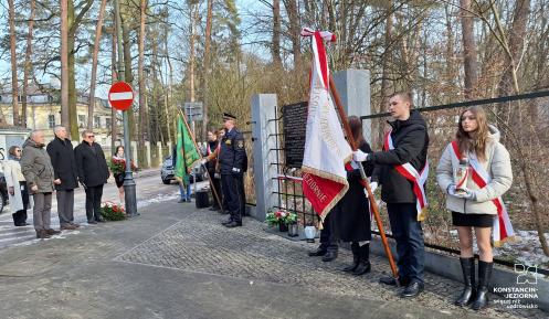 Grupa młodych dziewczyn i chłopców z biało-czerwonymi wieńcami i szarfami oraz trzech starszych mężczyzn i trzy kobiety stoją naprzeciwko tablicy zawieszonej na ogrodzeniu. Przy ogrodzeniu dwóch strażników miejskich w galowych mundurach.