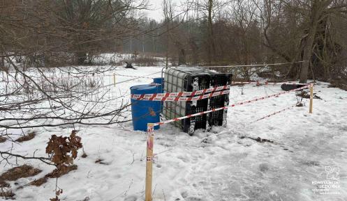 Na zaśnieżonym terenie przy zadrzewionej drodze stoją zabezpieczone taśmą ostrzegawczą pojemniki – mauzer i dwie beczki – odgrodzone drewnianymi palikami.