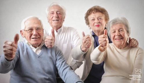 Czworo uśmiechniętych seniorów siedzi obok siebie i pokazuje uniesione kciuki na znak zadowolenia.