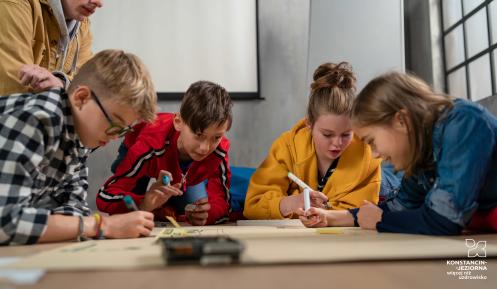Grupa dzieci siedzi przy stole i wspólnie rysuje na dużych arkuszach papieru, korzystając z kolorowych markerów, pod opieką dorosłego opiekuna w sali zajęć.