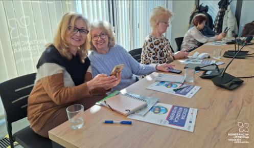 Dwie kobiety siedzą przy stole, patrzą w obietkyw i uśmiechają się. W rękach trzymają telefony komórkowe, przed nimi na stole leżą broszury, zeszyty - są na szkoleniu.