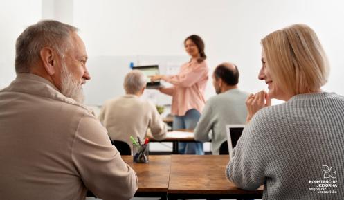Widok zza pleców pary seniorów siedzących przy biurku w sali wykładowej. Mężczyzna z brodą i kobieta o jasnych włosach uśmiechają się do siebie. Na pierwszym planie widać organizer z przyborami do pisania. W głębi sali, na rozmytym tle, inna uczestniczka prowadzi prezentację.
