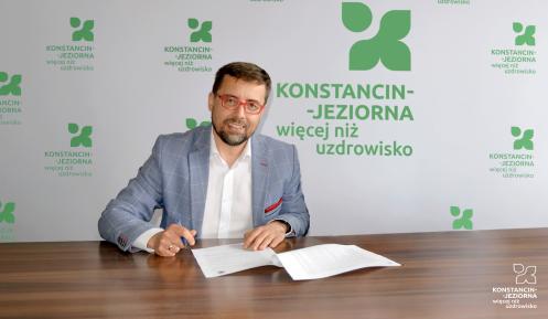 Mężczyzna w okularach podpisuje dokumenty przy stole na tle ścianki z logo „Konstancin-Jeziorna – więcej niż uzdrowisko”.