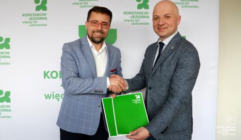 Dwóch mężczyzn w garniturach uściskiem dłoni potwierdza podpisanie dokumentu na tle ścianki z logo Konstancina-Jeziorny.