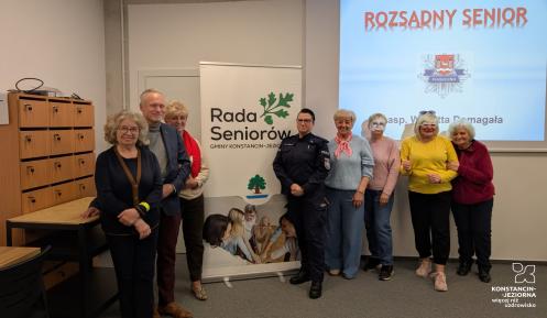 Grópa seniorów stoi w sali przy roll-upie z napisem Rada Seniorów, w tle na ścianie slajd prezentacji Rozsądny Senior