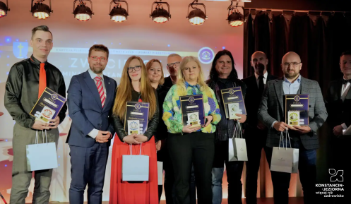 Grupowe zdjęcie dziesięciu osób na podświetlonej scenie, uchwycone podczas ceremonii wręczania nagród „Perła Mazowsza 2024”. W pierwszym rzędzie laureaci trzymają dyplomy i papierowe torby z nagrodami.