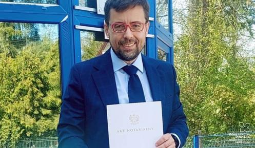 Mężczyzna w garniturze stoi przed budynkiem i trzyma w rękach dokument zatytułowany „Akt notarialny”.