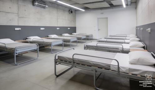 Nowoczesne pomieszczenie schronowe z betonowymi ścianami i oświetleniem LED, w którym ustawiono rzędy metalowych łóżek z materacami i poduszkami.