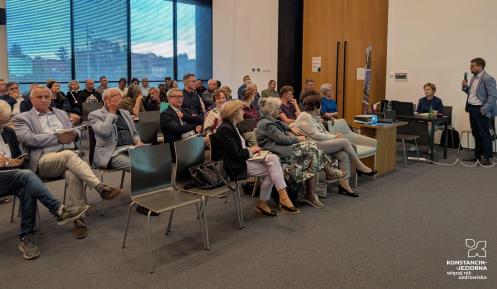 Uczestnicy spotkania siedzą na sali konferencyjnej i słuchają mężczyzny prezentującego temat przy użyciu mikrofonu i projektora.