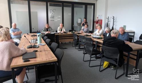 Fotografia przedstawia posiedzenie Rady Seniorów Gminy Konstancin-Jeziorna w nowoczesnej sali konferencyjnej. Grupa kilkunastu osób siedzi przy stołach ułożonych w kształt litery U. Na pierwszym planie po lewej widać tył sylwetki kobiety w różowym swetrze, a przed nią laptop i mikrofon konferencyjny na drewnianym blacie. Pozostali uczestnicy obrad, w większości seniorzy, siedzą przodem lub bokiem do obiektywu, słuchając wystąpienia. W głębi sali, za szklaną ścianą z drzwiami, na której widnieje logo gminy (