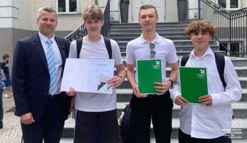 Na schodach przed budynkiem stoją cztery osoby: zastępca burmistrza gminy oraz trzej uczniowie trzymający dyplom i zielone teczki z nagrodami, laureaci konkursu wiedzy o historii Konstancina-Jeziorny.