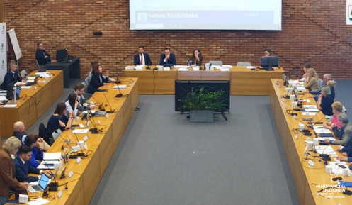 Na zdjęciu sala obrad Rady Miejskiej Konstancin-Jeziorna podczas sesji – radni siedzą przy stołach ustawionych w kształcie podkowy, a obrady prowadzi prezydium zasiadające przy stole przewodniczącym.