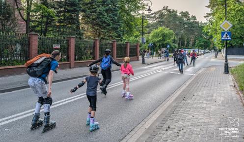 Grupa dorosłych i dzieci w kaskach oraz ochraniaczach jedzie na rolkach środkiem asfaltowej ulicy. W tle widać drzewa, murowane ogrodzenie, chodnik oraz innych uczestników rolkowego przejazdu.