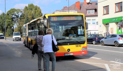 Żółto-czerwony autobus linii 710 zatrzymuje się na przystanku w centrum Konstancina-Jeziorny, gdzie na chodniku czekają pasażerowie.