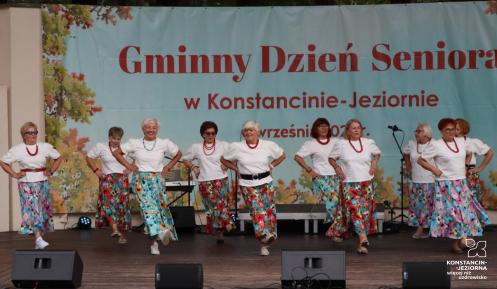 Grupa seniorek w jednakowych białych bluzkach, czerwonych koralach i kolorowych, wzorzystych spódnicach tańczy na scenie amfiteatru. W tle widać duży baner z napisem „Gminny Dzień Seniora w Konstancinie-Jeziornie”.