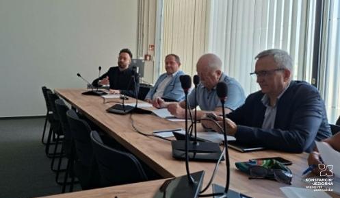Fotografia przedstawia posiedzenie Rady Seniorów Gminy Konstancin-Jeziorna w nowoczesnej sali konferencyjnej. Grupa kilkunastu osób siedzi przy stołach ułożonych w kształt litery U. Na pierwszym planie po lewej widać tył sylwetki kobiety w różowym swetrze, a przed nią laptop i mikrofon konferencyjny na drewnianym blacie. Pozostali uczestnicy obrad, w większości seniorzy, siedzą przodem lub bokiem do obiektywu, słuchając wystąpienia. W głębi sali, za szklaną ścianą z drzwiami, na której widnieje logo gminy (
